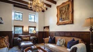 Ein geräumiges Wohnzimmer mit hohen Decken, Holzvertäfelung, einer großen Chandelierleuchtre, einem Sofa mit vielen Kissen, einem Couchtisch und einem Gemälde an der Wand.