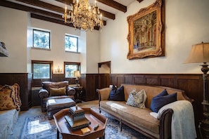 Ein geräumiges Wohnzimmer mit hohen Decken, Holzvertäfelung, einer großen Chandelierleuchtre, einem Sofa mit vielen Kissen, einem Couchtisch und einem Gemälde an der Wand.