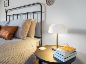 A neatly made bed with a headboard, a bedside table with a lamp and books, and a wall with a decorative plate. - Rosamund Townhouse Clifden (Clifden)