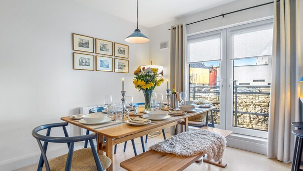 Een eetruimte met een houten tafel voor vier personen, blauwe stoelen, een bank, een vaas met bloemen en lijstjes met foto's aan de muur.