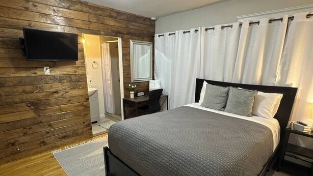 A modern bedroom with a wooden wall, a bed with grey bedding, a bedside table, a TV mounted on the wall, and a ceiling light. - Private Suite w/ Bath & Free Parking (Brockton)
