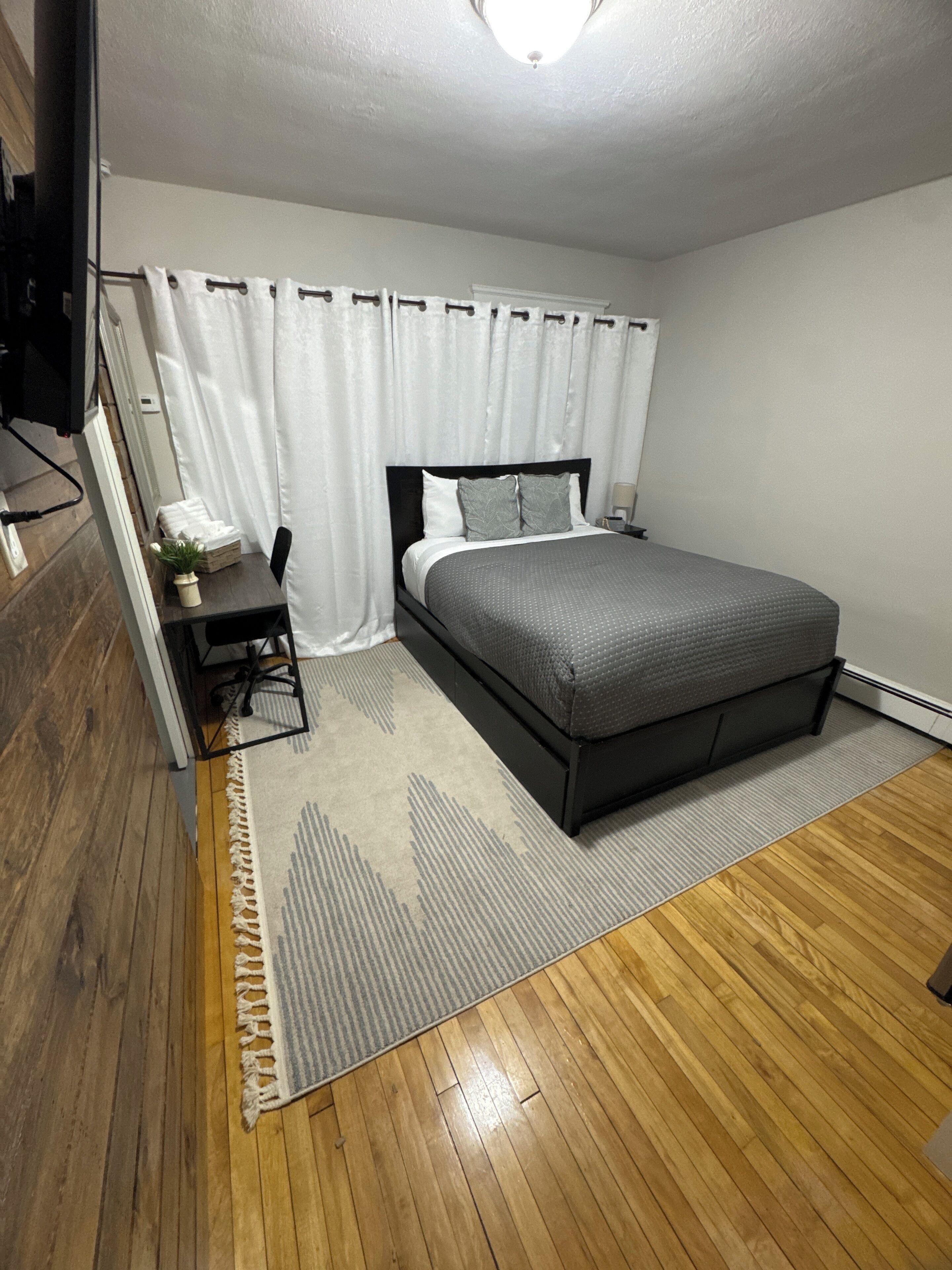 A bedroom with a bed, a desk, a chair, a small table with a plant, and a TV.