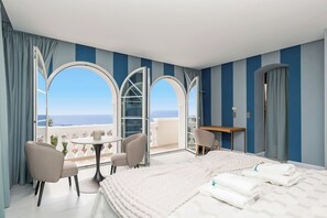 Ein Hotelzimmer mit einem großen Bett, zwei Sesseln, einem kleinen runden Tisch und einem Schreibtisch. Große Fenster bieten Blick aufs Meer.