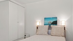 Een slaapkamer met een bed, twee kussens, een nachtkastje, een kledingkast en een schilderij van een kustlandschap.