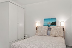Ein Schlafzimmer mit Bett, zwei Kissen, einem Nachttisch, einem Kleiderschrank und einem Gemälde einer Küstenlandschaft.