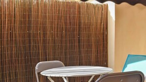 Een klein terras buiten met een ronde tafel en twee stoelen, een bamboescherm en een gedeeltelijk uitzicht op een gebouw.