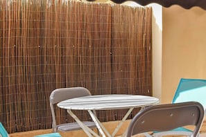 Eine kleine Terrasse im Freien mit einem runden Tisch, zwei Stühlen, einem Bambuszaun und einem teilweisen Blick auf ein Gebäude.