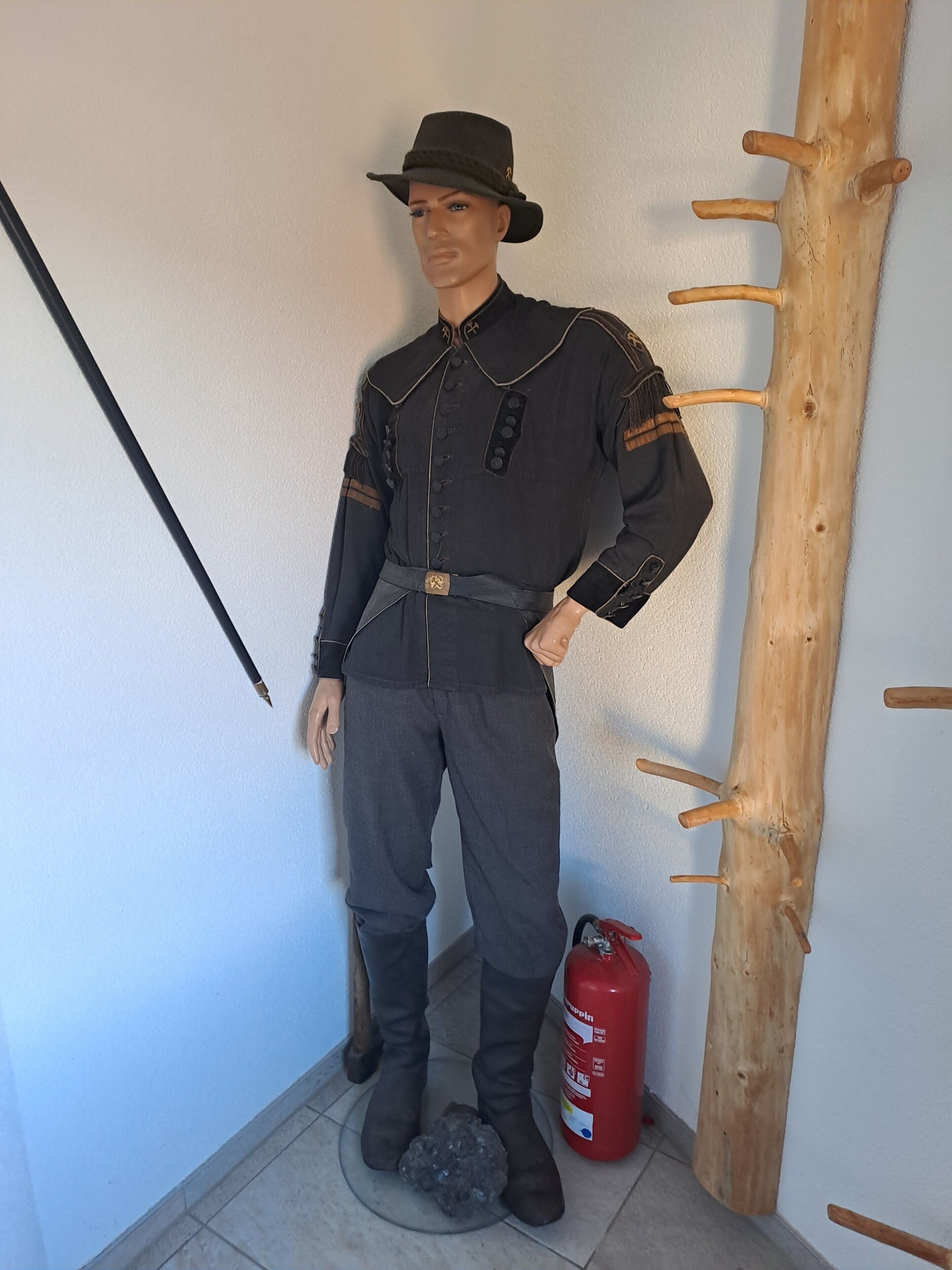 En dukke iført mørk uniform med bælte står ved siden af et træbeskyttet opslagstavle og en rød brandslukker.