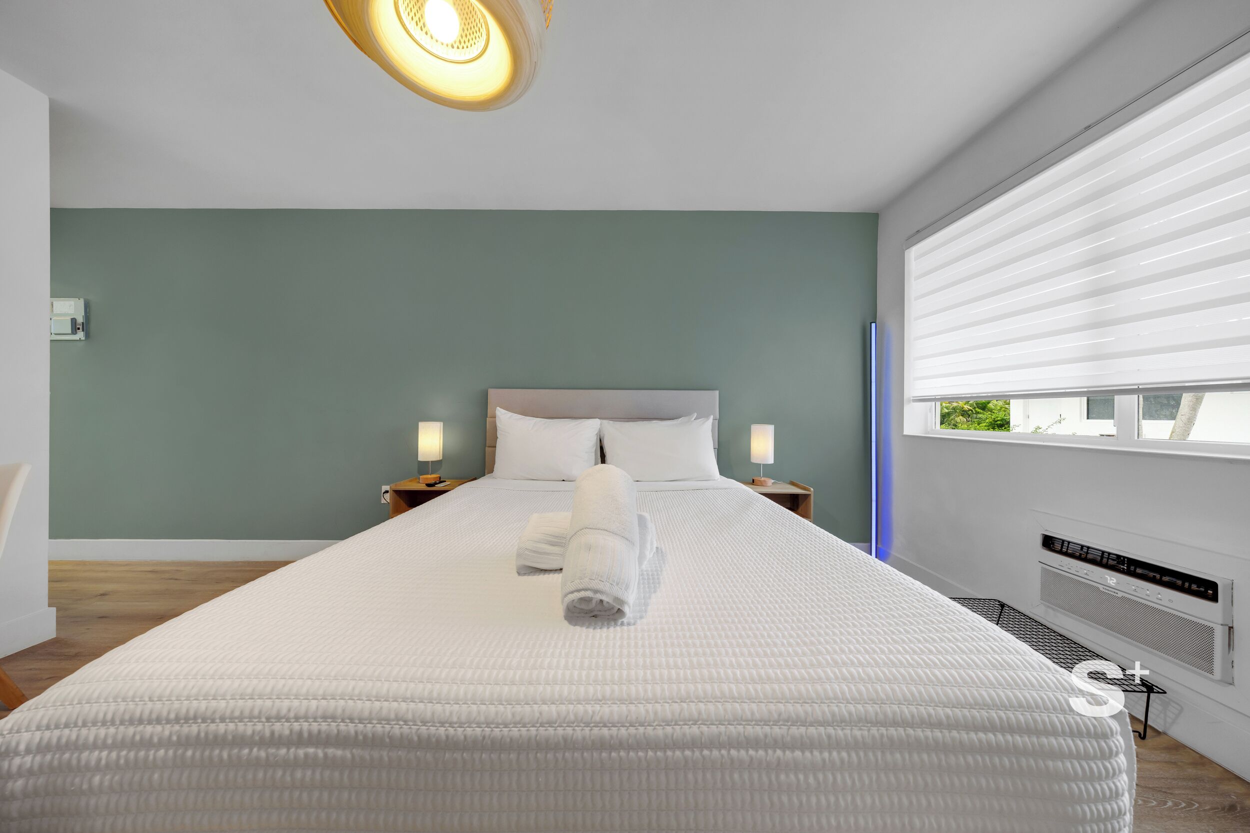 A modern hotel room with a large bed, two bedside lamps, a window with blinds, and a ceiling light fixture.
