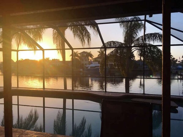 Blick durch ein Fenster mit Metallrahmen, das einen Sonnenuntergang über Wasser mit Palmen und Gebäuden im Hintergrund reflektiert.
