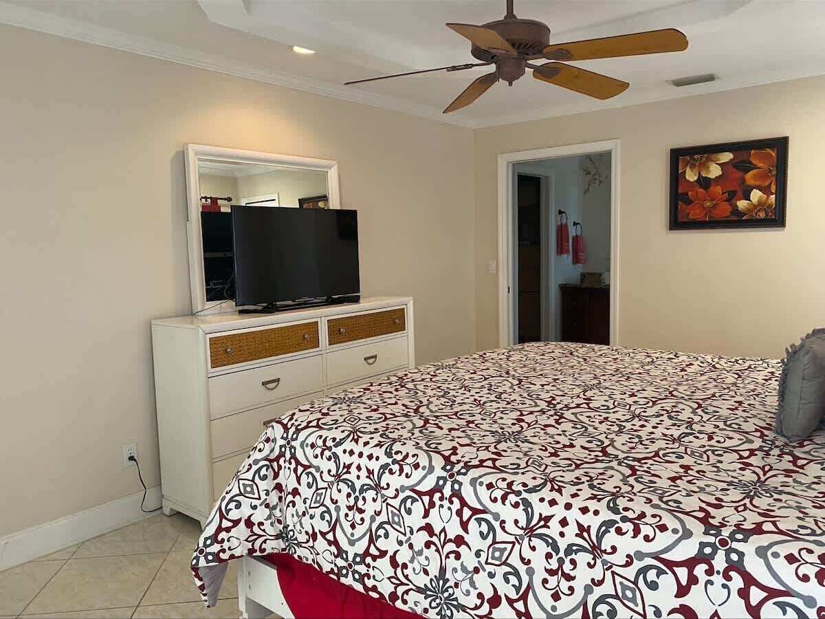 A bedroom with a bed, a dresser, a television on the dresser, a ceiling fan, and a door leading to another room.