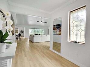 Ein geräumiges Zimmer mit Holzboden, einem weißen Sofa, einem Esstisch mit Stühlen und einem großen Fenster mit geometrischem Muster.