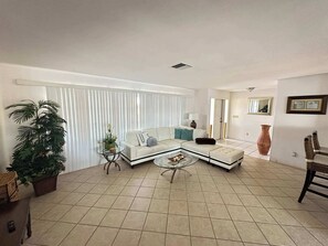 Ein geräumiges Wohnzimmer mit einem Ecksofa, einem Glastisch und einer großen Kübelpflanze.