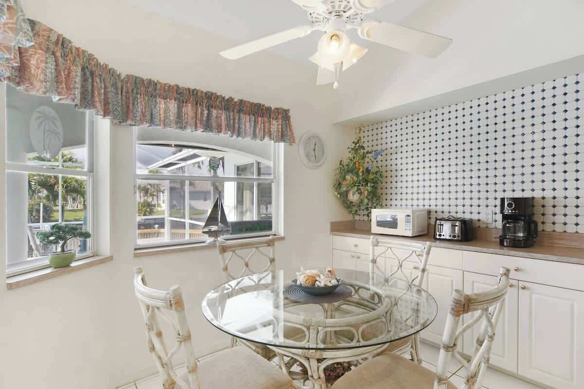 A dining area with a glass table and white chairs, a ceiling fan, a window with a view, and a kitchen counter with appliances.