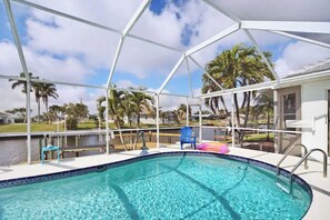 Ein Swimmingpool mit weißer Liegefläche und überdachtem Bereich, umgeben von Palmen und Blick auf ein Gewässer.