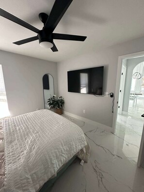 Ein Schlafzimmer mit Bett, Deckenventilator, Fernseher, Topfpflanze und einem Badezimmer mit Glastür.