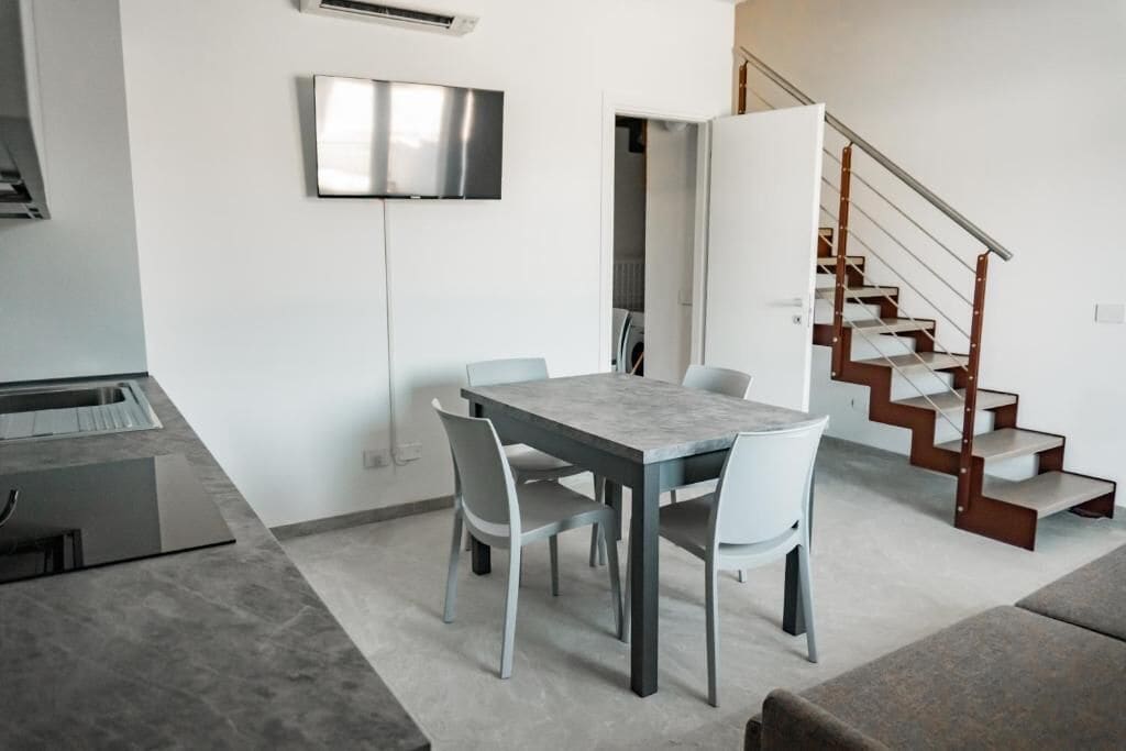 A modern kitchen with a built-in stove, a dining area with a marble table and white chairs, and a staircase with wooden railings.