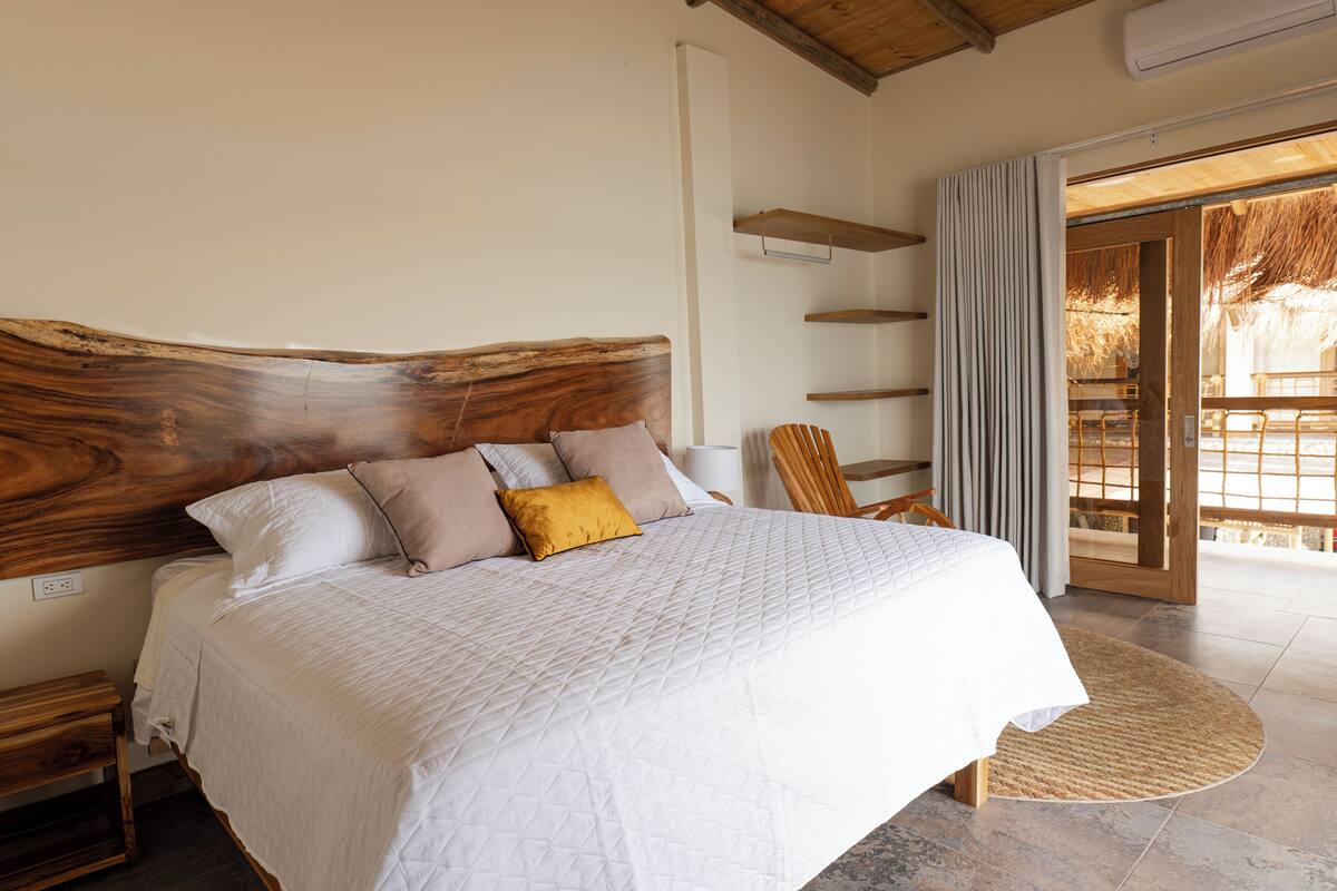A bedroom with a large bed, a wooden headboard, a bedside table, a chair, and a view of the outside.