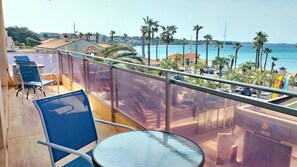 A balcony with a table and chairs, overlooking a beach with palm trees and a clear sky. - Panoramic Sea View and Beaches on Foot (Six-Fours-les-Plages)