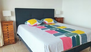 A bed with a dark headboard, white bedding, and colorful geometric-patterned pillows. There are two bedside tables with lamps and a wooden dresser with drawers. - Panoramic Sea View and Beaches on Foot (Six-Fours-les-Plages)