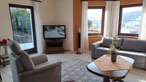 Ein Wohnzimmer mit Sofa, Sessel, Fernseher und Blick auf eine verschneite Landschaft durch das Fenster.