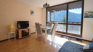 Ein modernes Wohnzimmer mit Fernseher, Esstisch, Stühlen und einem großen Fenster mit Bergblick.