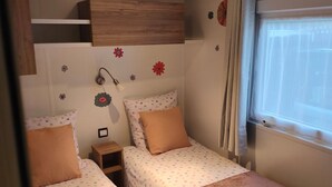 A compact room with two beds, a small bedside table, a wall-mounted shelf, and a window with curtains. - Mobile Home “Lurielle” with Shared Pool, Communal Terrace, and Private Garden (Blonville-sur-Mer)