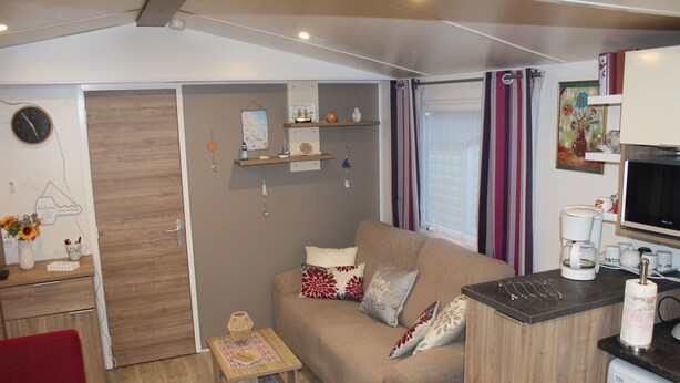 A compact living space with a sofa, a small table, a washing machine, and a kitchenette area. - Mobile Home “Lurielle” with Shared Pool, Communal Terrace, and Private Garden (Blonville-sur-Mer)
