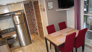A compact kitchen with a refrigerator, stove, and wooden flooring. - Mobile Home “Lurielle” with Shared Pool, Communal Terrace, and Private Garden (Blonville-sur-Mer)