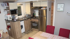 A compact kitchen with a microwave, stove, and refrigerator; a dining area with a table and chairs; and a washing machine in the corner. - Mobile Home “Lurielle” with Shared Pool, Communal Terrace, and Private Garden (Blonville-sur-Mer)