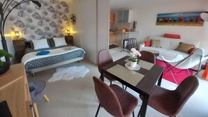 Un dormitorio moderno con cama, mesitas de noche y una mesilla. Se observa un comedor con mesa, sillas y un jarrón con flores. También se distingue una sala de estar con sofá y un cuadro.