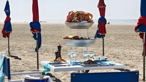 Mobiliario de comedor junto a la playa, con una parrilla para mariscos, mesa con platos, vasos y sombrillas.