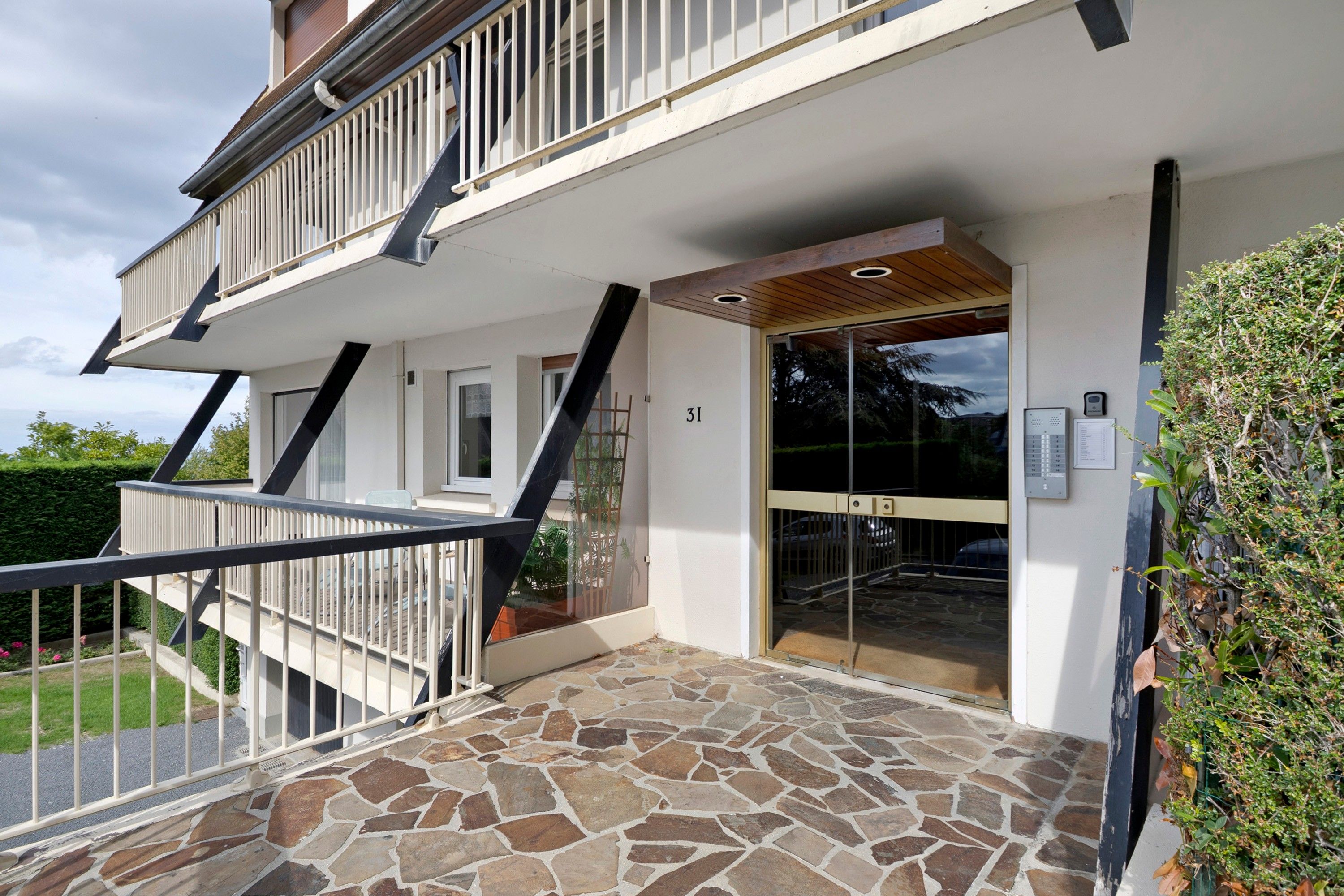Een gebouw met twee verdiepingen, een entree met stenen tegels, een balkon met metalen hekwerk en een houten plafond.