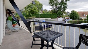 En balkon med to sorte vævede borde og stole, gelænder i hvidt og udsigt til gaden samt grønne områder.
