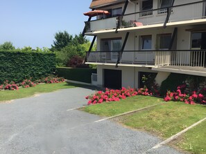 Un immeuble à plusieurs étages doté de balcons, d’une allée pavée et d’un jardin bien entretenu agrémenté de fleurs roses.