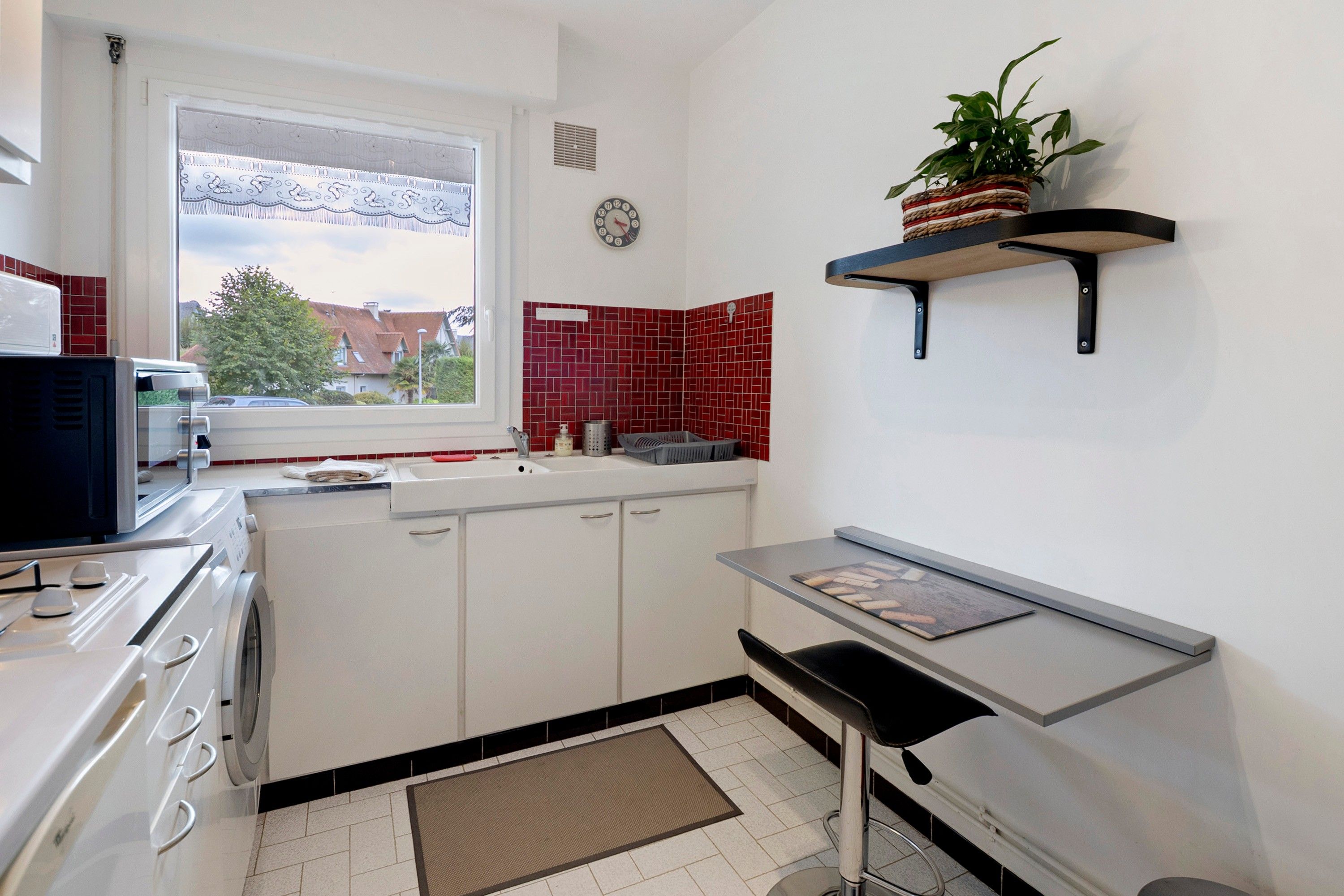 Een kleine keuken met witte kastjes, een ingebouwde magnetron, een spoelbak en een raam met uitzicht op huizen.