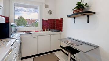 Een kleine keuken met witte kastjes, een ingebouwde magnetron, een spoelbak en een raam met uitzicht op huizen.