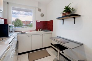 Une cuisine compacte avec des meubles blancs, un four à micro-ondes encastré, un évier et une fenêtre donnant sur des maisons.