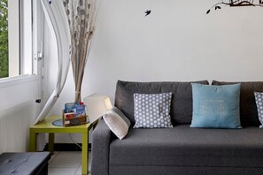 A modern living room with a grey sofa, patterned pillows, a small side table with a lamp, and a window with a view of trees.