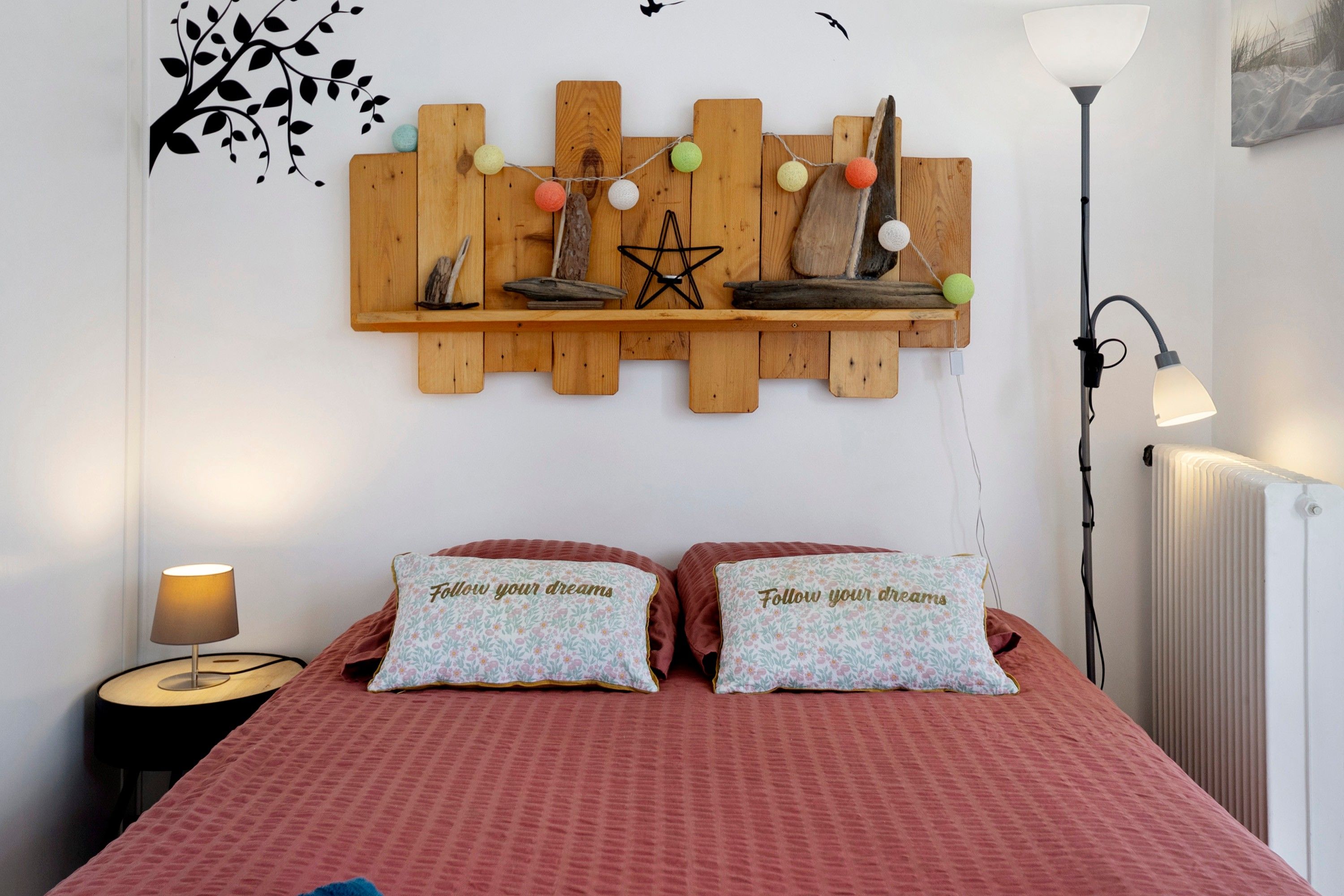 Ein Schlafzimmer mit einem Bett, Kissen, einem Nachttisch, einer Wandablage mit Dekoration und einer Stehlampe.