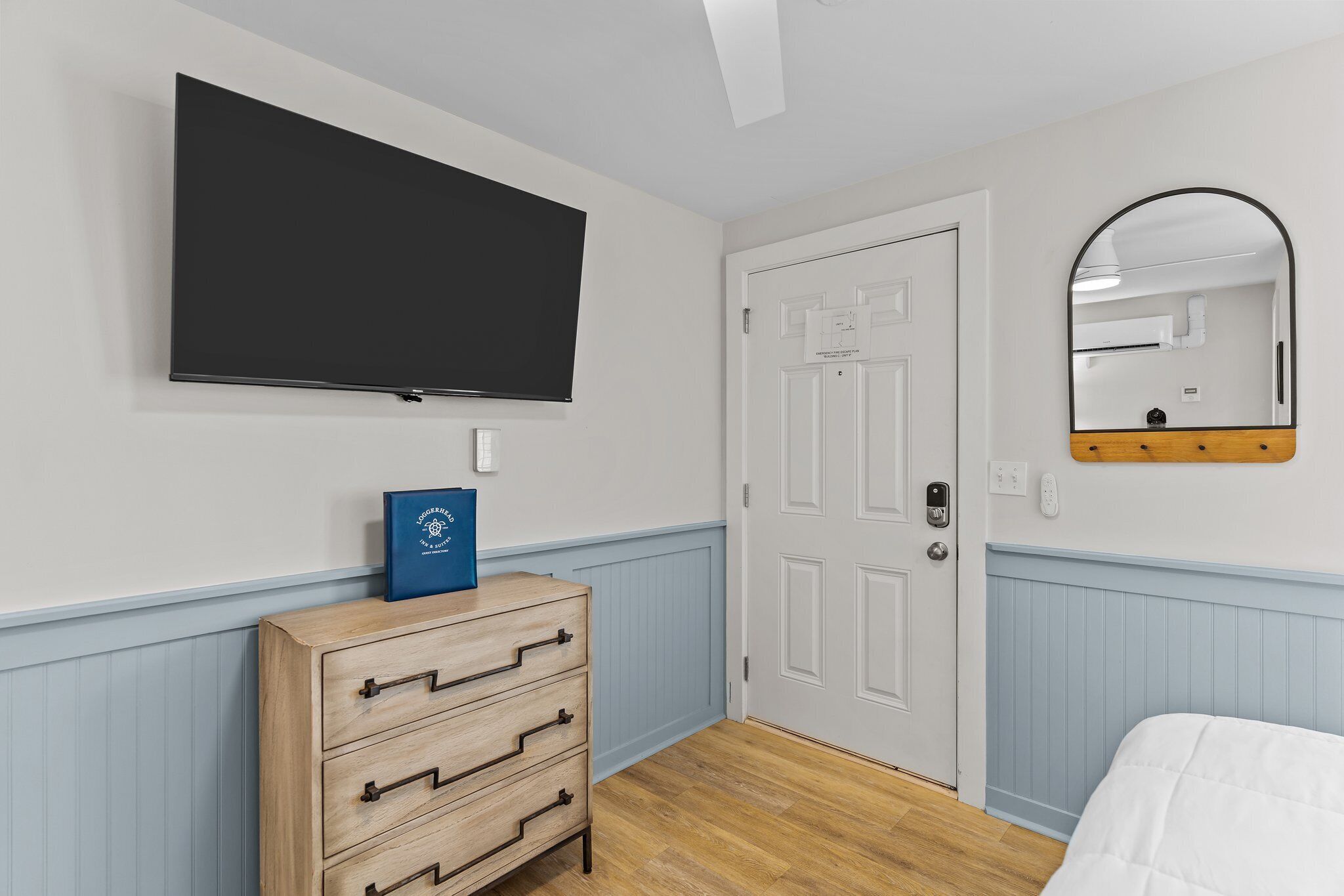 Een slaapkamer met een flatscreen-tv aan de muur, een houten dressoir met laden, een witte deur en een spiegel met houten lijst.