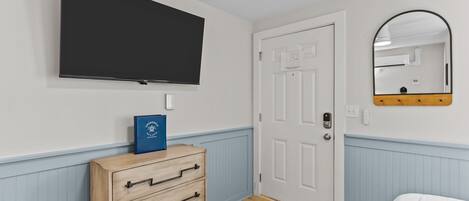 A bedroom with a flat-screen TV mounted on the wall, a wooden dresser with drawers, a white door, and a mirror with a wooden frame.