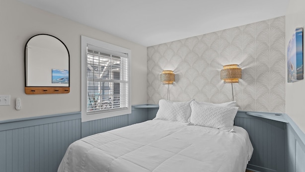 Een slaapkamer met een bed, een raam met zonwering, een spiegel aan de muur en twee wandlampen.