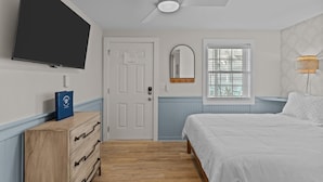 Een slaapkamer met een bed, een plafondventilator, een televisie, een dressoir, een spiegel en een raam.