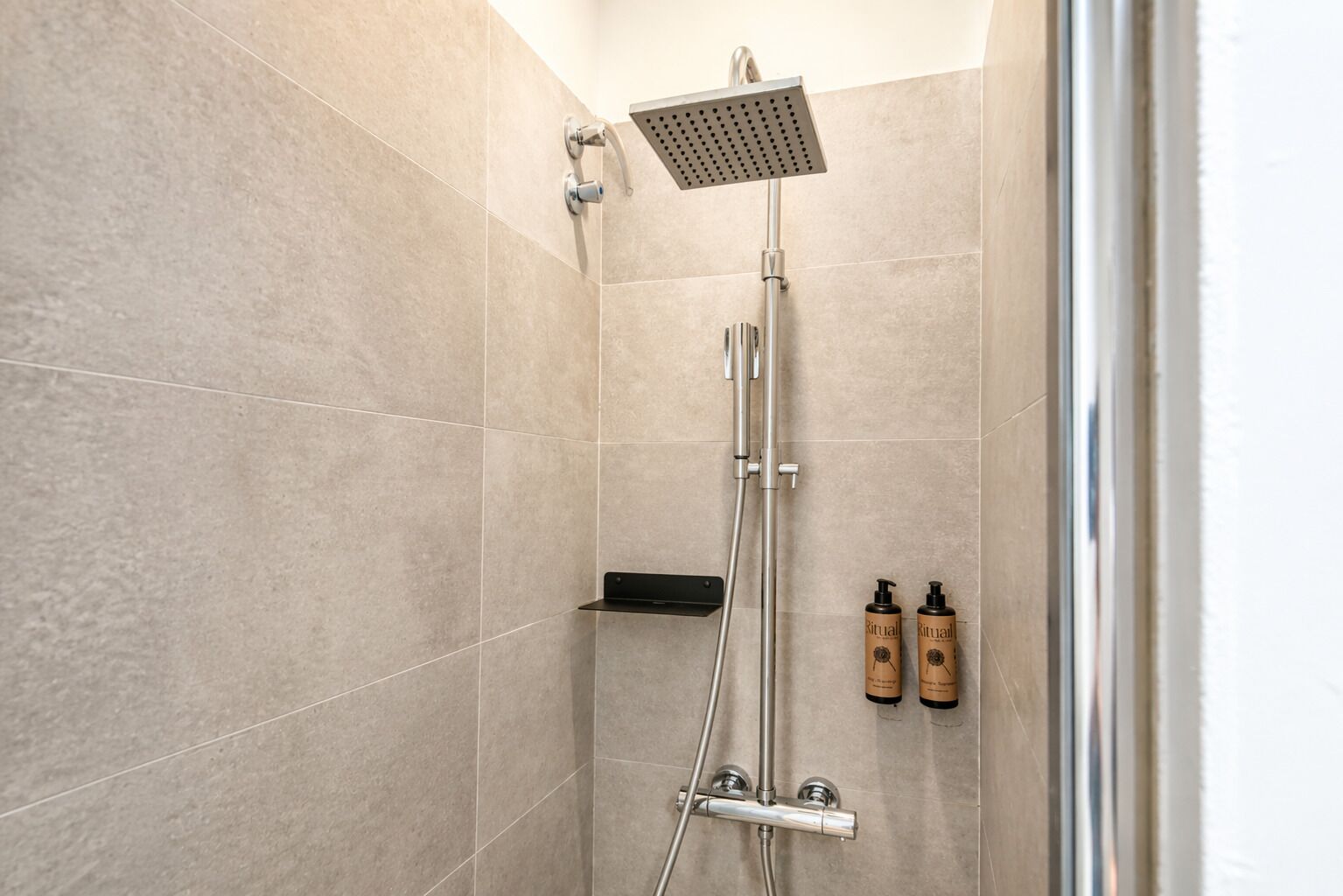 Une douche équipée d’un pommeau de douche à main, d’un pommeau de douche fixé au mur et de deux flacons de gel douche Ritual placés sur une étagère.