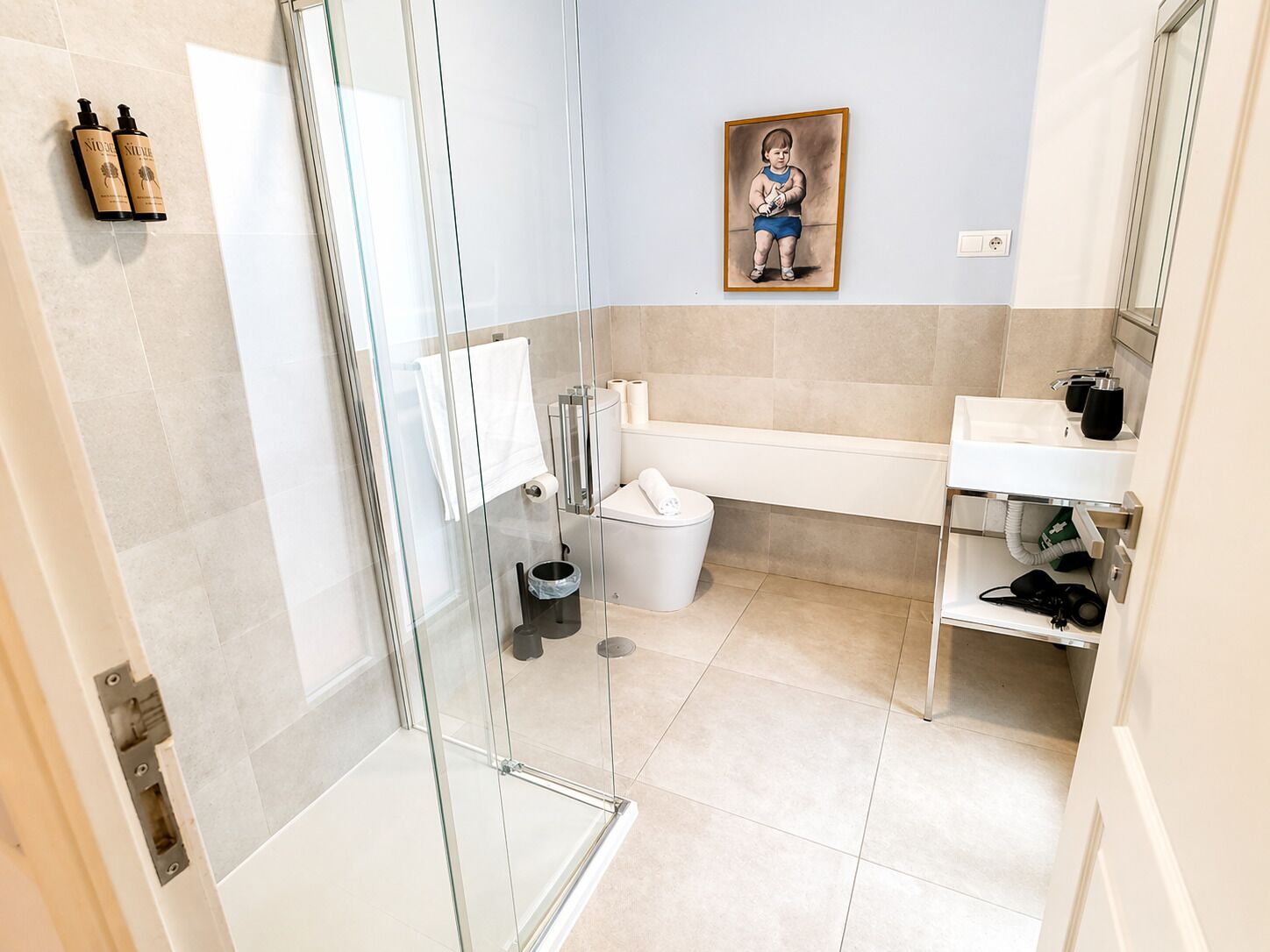Ein Badezimmer mit einer Duschkabine aus Glas, einem weißen Waschbecken, einer Toilette und einem gerahmten Bild an der Wand.