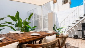 A wooden dining table set with plates and glasses, surrounded by wicker chairs, with a staircase and outdoor grill in the background. - Prado Suites Apartments (Málaga)