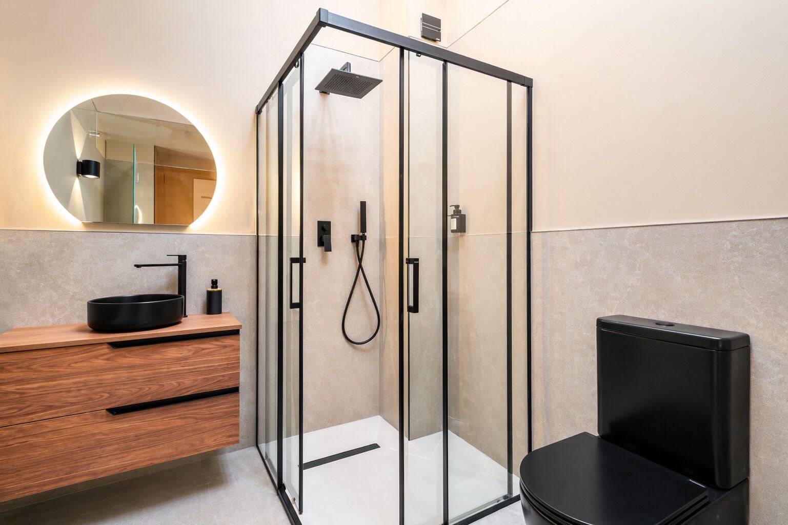 Un baño moderno con cabina de ducha de vidrio, un espejo redondo, tocador de madera y una silla negra.