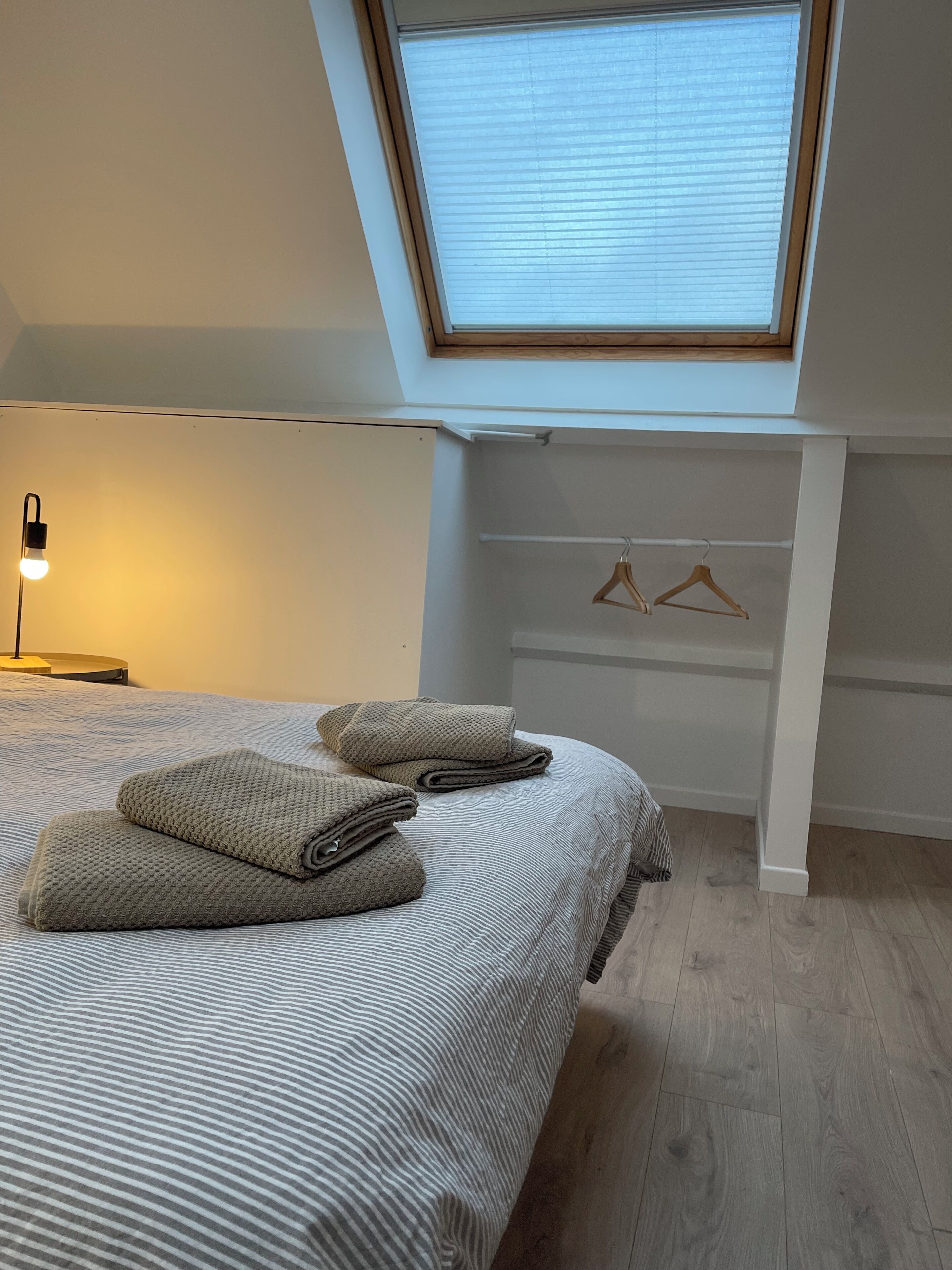 A bedroom with a skylight, a bed with striped bedding, two pillows, a bedside lamp, and a wooden floor.