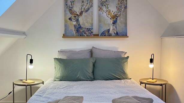 Quarto com teto inclinado, cama com roupa de cama listrada, duas almofadas, duas mesas de cabeceira com abajures e duas pinturas de veados na parede.